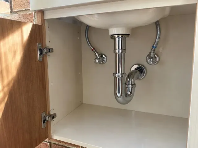 Open vanity cabinet under a sink with neat chrome drain pipes and dry white walls in Glenhaven